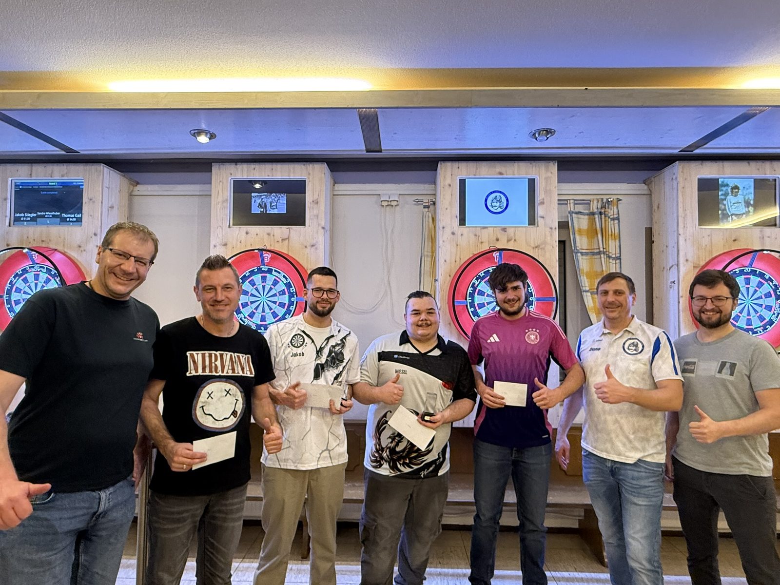 Siegerehrung des Neujahrs-Darts-Turniers 2026: Teilnehmer mit Preisen und erhobenem Daumen vor den beleuchteten Boards im Sportheim Niederhofen