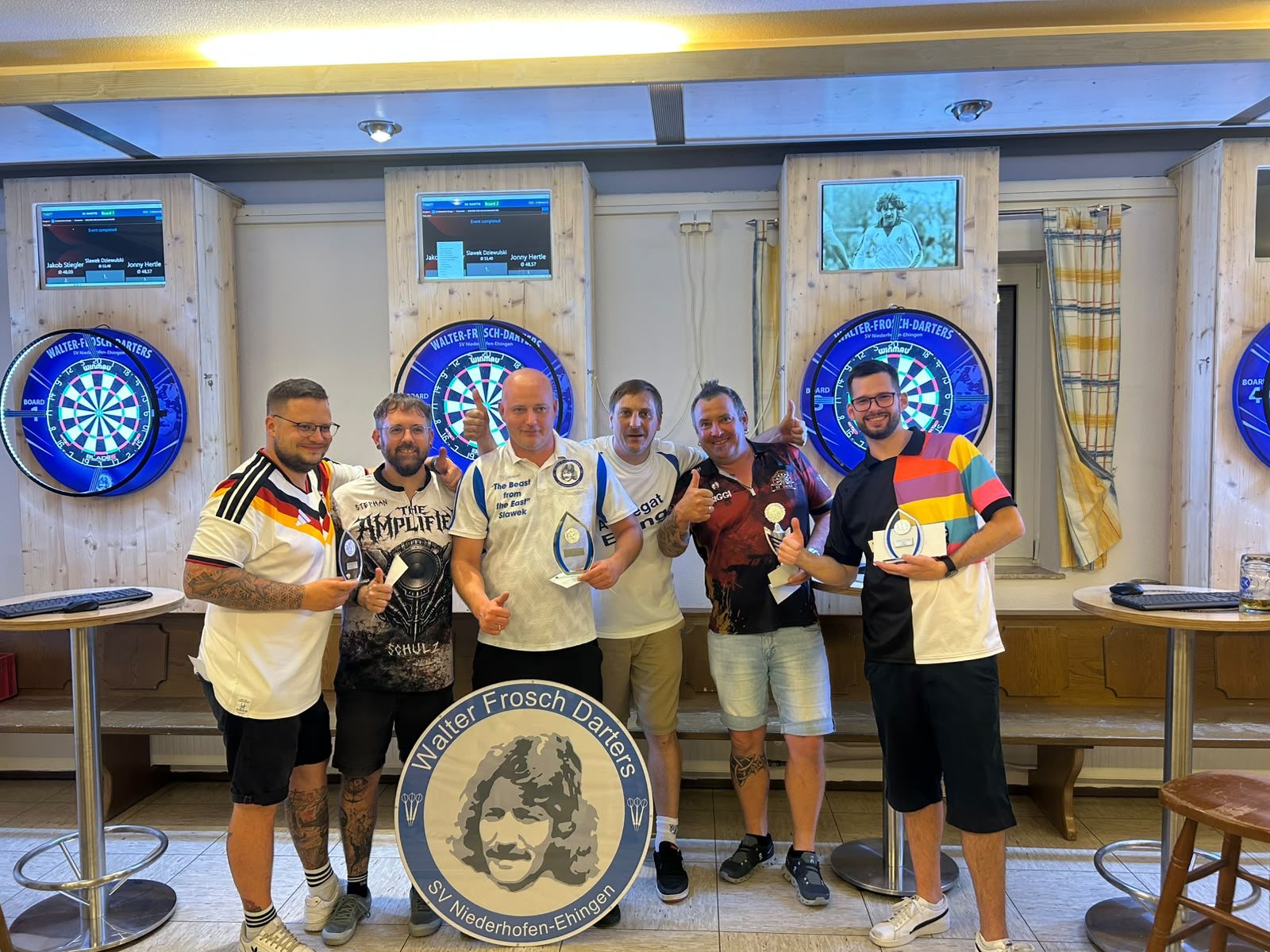 Sechs Dartspieler mit Pokalen vor den Dartscheiben im Sportheim
