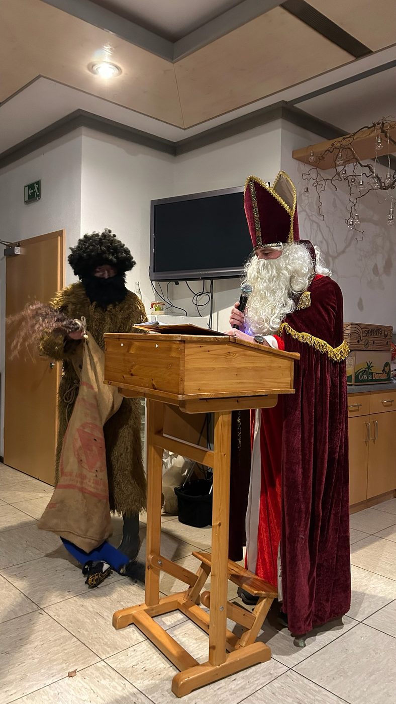 Nikolaus mit Mikrofon am Rednerpult und Knecht Ruprecht mit Jutesack bei der Weihnachtsfeier im Sportheim